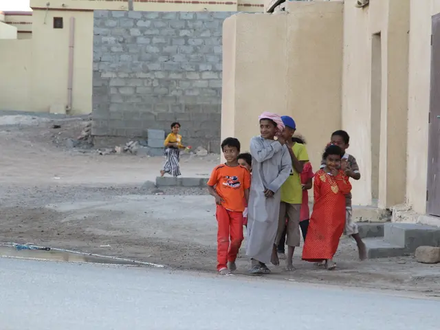In this image there are group of kids standing and smiling , and there are buildings, water, rocks,...